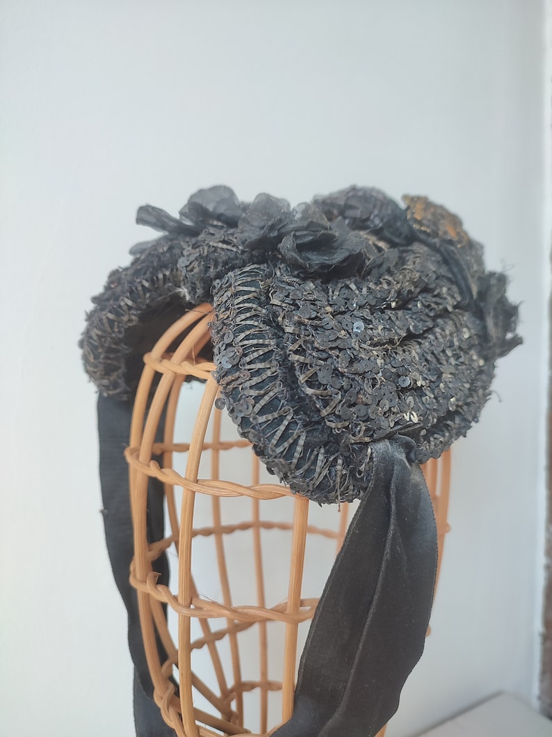 May include: A black sequined hat with a large black bow and floral embellishments. The hat is displayed on a wooden hat form.