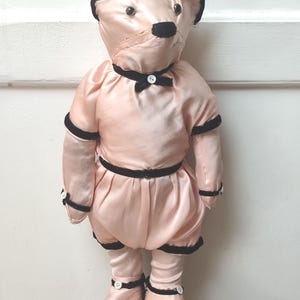 Peut inclure: Une poupée ours en peluche vintage vêtue d'une tenue en satin rose avec des bordures noires. L'ours a un nez noir, des oreilles noires et un petit nœud papillon noir. La tenue de la poupée comprend une jupe et des bottes assorties avec des boutons blancs.