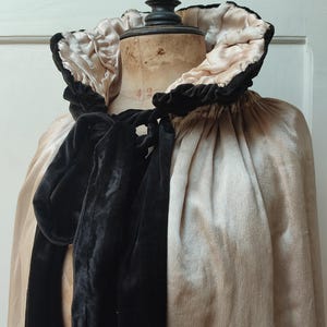 May include: A vintage cape in a soft, champagne color, featuring a large, ruffled collar with black velvet trim. A black velvet bow detail adorns the front. The cape is draped over a mannequin.