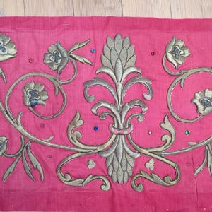 Op de afbeelding: Een stuk roze stof met sierlijke gouden en zilveren borduursels. Het ontwerp toont gestileerde bloemen, bladeren en een centraal symmetrisch motief. Kleine, kleurrijke kralen zijn over het oppervlak verspreid en voegen een vleugje glans toe.