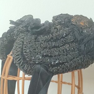 May include: A black sequined hat with a black ribbon and a decorative gold embellishment. The hat is displayed on a wooden stand.