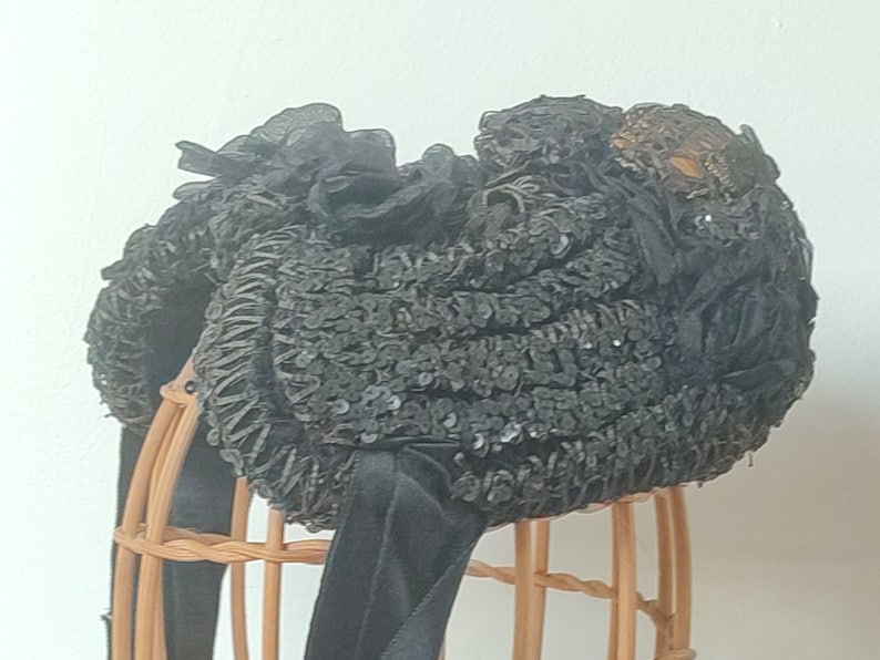 May include: A black sequined hat with a black ribbon and a decorative gold embellishment. The hat is displayed on a wooden stand.