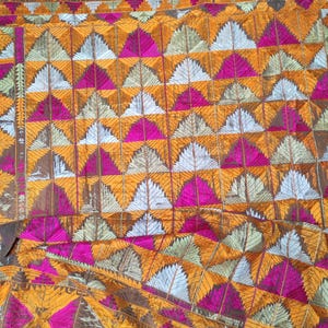 Peut inclure: Gros plan sur un textile avec un motif géométrique répétitif. Le motif présente des triangles et des losanges dans des tons or, rose, bleu clair et vert, avec une bordure marron. Le tissu semble être brodé ou tissé.