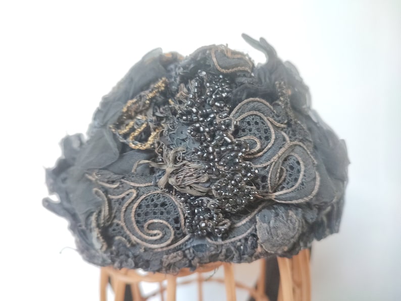 May include: A black vintage hat with intricate lace detailing and black beads. The hat is adorned with gold embroidery and has a slightly ruffled texture.