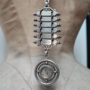 Puede incluir: Colgante plateado con una brújula en la parte inferior y una serie de paneles rectangulares grabados. El colgante está suspendido de una cadena.