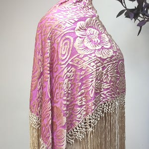 May include: A vibrant pink shawl featuring a raised, textured beige floral and swirl pattern in velvet. The garment is finished with long, delicate beige fringe along the edges, creating a layered look with a soft, tactile surface throughout.