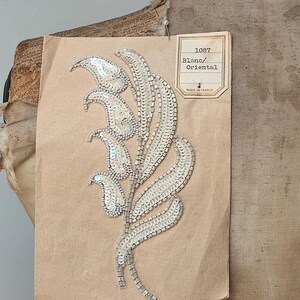 Peut inclure: Applique florale blanche à paillettes sur une étiquette en papier brun. L'applique est composée de paillettes et de perles irisées, avec un motif de tige et de feuilles. L'étiquette indique "1087 Blanc/Oriental Made in France."