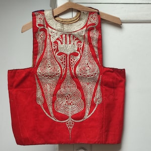 Peut inclure: Un gilet rouge orné de broderies argentées complexes, suspendu à un cintre en bois. La broderie présente un motif symétrique avec des motifs floraux et géométriques. Le gilet a une encolure arrondie et une forme rectangulaire.