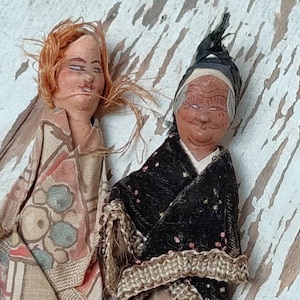 Könnte beinhalten: Zwei antike Puppen mit bemalten Gesichtern und Stoffkleidung. Die Puppe links hat rote Haare und ein Kleid mit Blumenmuster. Die Puppe rechts hat ein schwarzes Samtkleid mit einer Häkelborte.