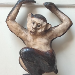 May include: A ceramic monkey sculpture with arms raised. The monkey has a light-colored face and body with darker brown accents on the head, arms, and legs. The sculpture has a distressed, aged appearance.