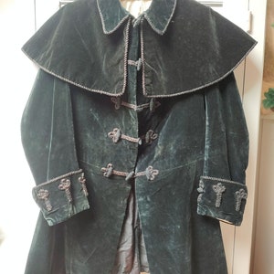 Puede incluir: Una chaqueta de terciopelo verde oscuro con un cuello grande y adornos decorativos. La chaqueta tiene un estilo largo y fluido y presenta una serie de botones en la parte delantera.