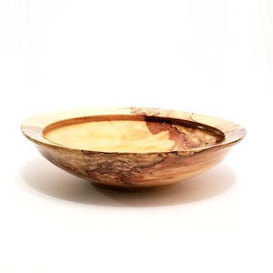 May include: A large, wooden bowl with a natural wood finish. The bowl has a wide, shallow shape and a slightly raised rim. The wood grain is visible and has a light brown color with darker brown streaks.