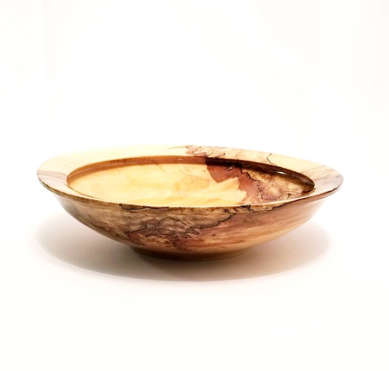 May include: A large, wooden bowl with a natural wood finish. The bowl has a wide, shallow shape and a slightly raised rim. The wood grain is visible and has a light brown color with darker brown streaks.