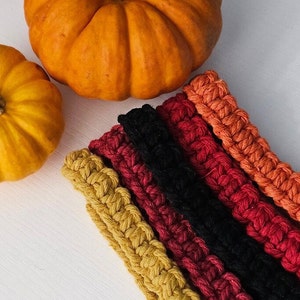 May include: An overhead shot features two pumpkins and several crocheted bands in fall colors. The pumpkins are orange and yellow. The bands are in shades of gold, red, black, and orange. The bands are arranged diagonally across the frame.