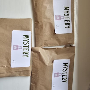 Puede incluir: Tres paquetes de papel marrón con etiquetas blancas que dicen "MYSTERY" en texto verde. Cada etiqueta tiene un pequeño gráfico de bolsa de compras rosa. Los paquetes están etiquetados con los precios £1, £2 y £3.