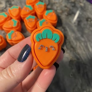 May include: Orange carrot-shaped charms with green tops and small gray facial features. The charms are in a pile, with one held up close. The charms are likely for crafting or jewelry making.