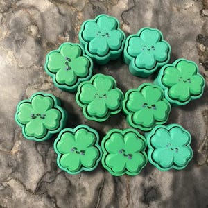 May include: A collection of ten green and light green clover-shaped objects with smiling faces and purple accents. The objects are arranged in a circular pattern, with a marbled background.