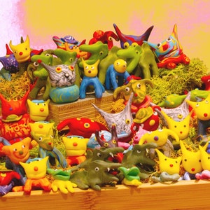 May include: A collection of colorful, whimsical, hand-painted ceramic figurines in various animal and creature shapes. The figurines are arranged on a wooden surface with a mossy green background.