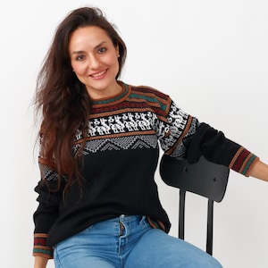 May include: A woman wearing a black sweater with a colorful geometric pattern and blue jeans. She is sitting on a black chair.