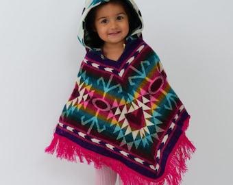 May include: A child wearing a colorful hooded poncho with a geometric pattern. The poncho has a fringe border and is made of a soft, knitted fabric.