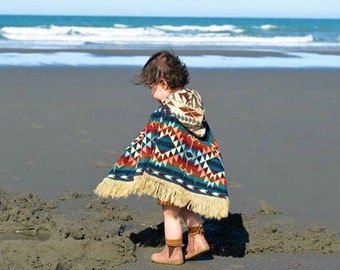 May include: A child wearing a colorful hooded poncho with a geometric pattern and fringe on the bottom. The poncho is blue, red, orange, yellow, and white. The child is standing on a sandy beach.