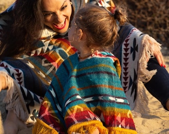 May include: A woman and a child are wearing colorful striped ponchos with fringe. They are sitting on a sandy beach and smiling at each other.
