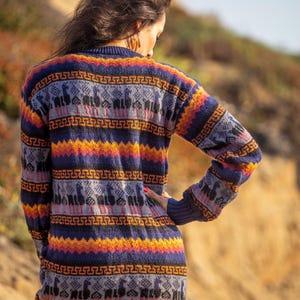 May include: A woman wearing a long, colorful sweater with a geometric pattern. The sweater features a repeating design of llamas and geometric shapes in shades of blue, orange, yellow, and gray.