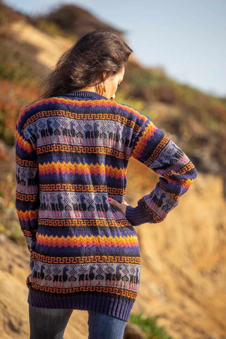 May include: A woman wearing a long, colorful sweater with a geometric pattern. The sweater features a repeating design of llamas and geometric shapes in shades of blue, orange, yellow, and gray.