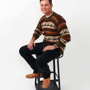 May include: A man wearing a brown, green, and white striped sweater with a geometric pattern. He is sitting on a black stool and wearing brown leather boots.
