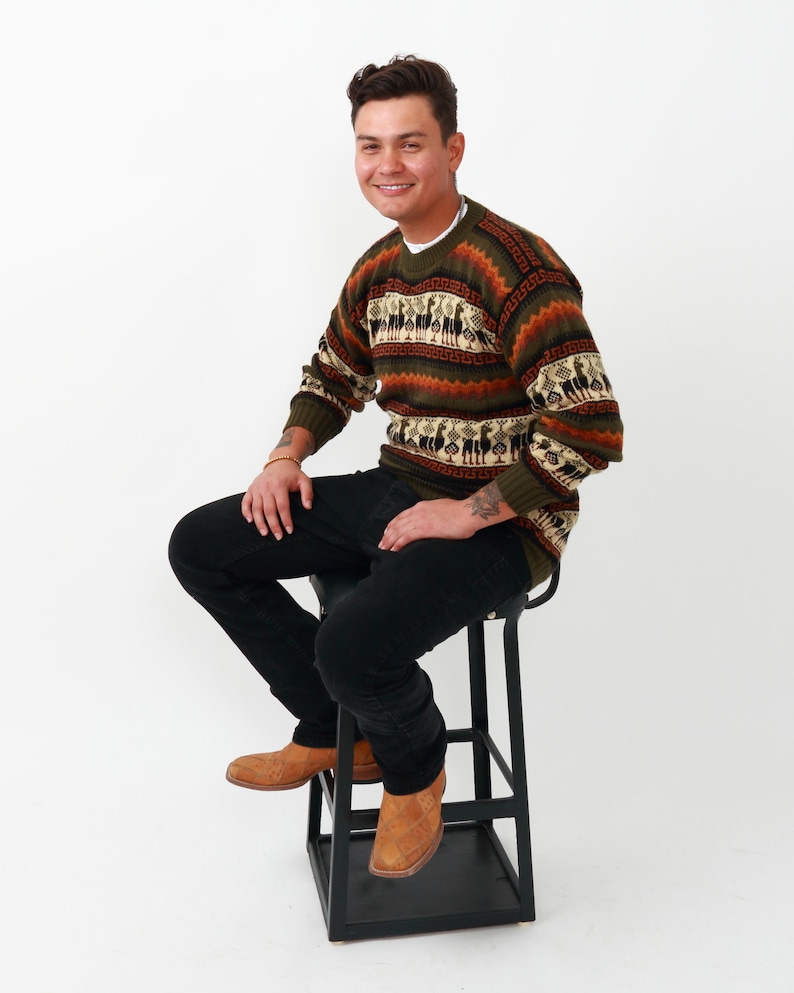 May include: A man wearing a brown, green, and white striped sweater with a geometric pattern. He is sitting on a black stool and wearing brown leather boots.