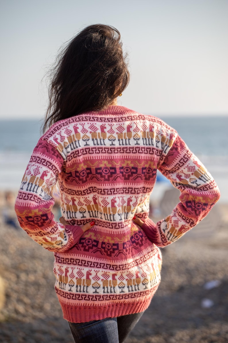May include: A woman wearing a pink and white sweater with a geometric pattern. The sweater features a repeating design of llamas and other geometric shapes.