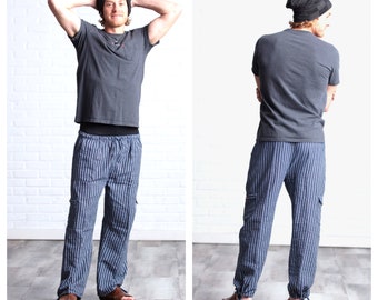 May include: A person wearing a gray t-shirt, a black beanie, and blue and white striped pants with a drawstring. The pants have cargo pockets on the sides. The person is standing in front of a white brick wall.