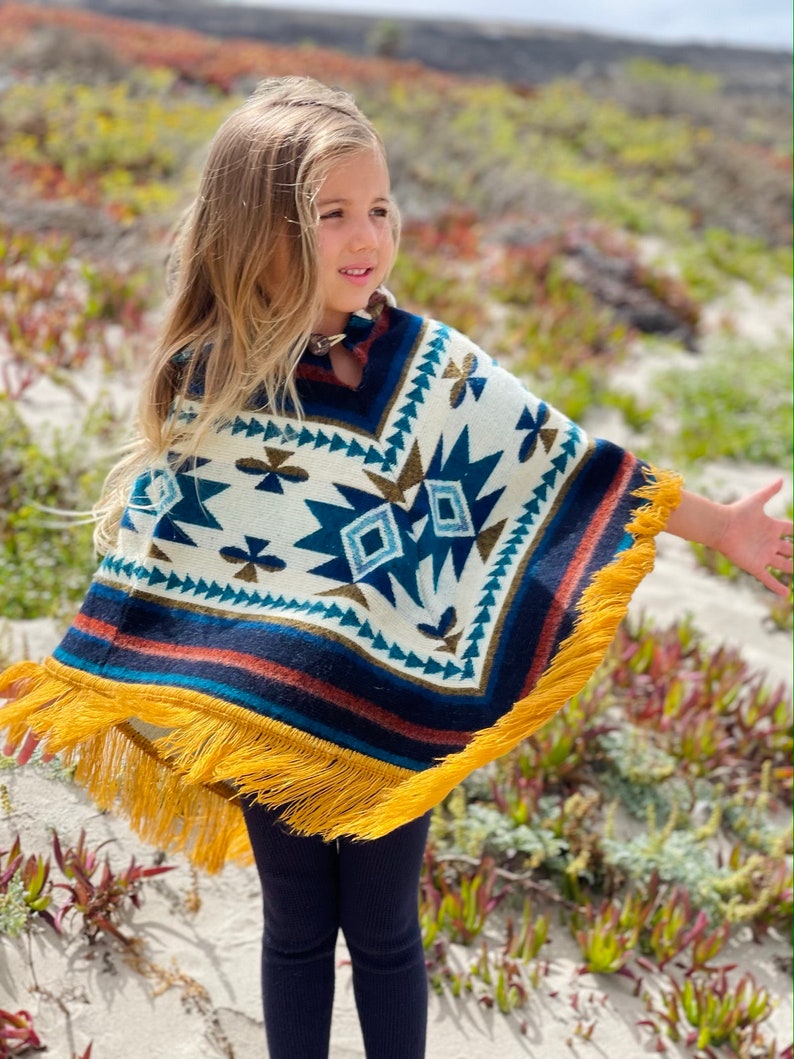 K&ouml;nnte beinhalten: Ein junges M&auml;dchen tr&auml;gt einen Poncho mit wei&szlig;en, blauen und braunen Mustern und einem gelben Fransenbesatz. Der Poncho hat ein geometrisches Muster und ist aus weichem, gestricktem Stoff gefertigt.