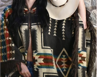 May include: A woman wearing a brown and black poncho with a geometric pattern and fringe. She is holding a white cowboy hat with a brown band.