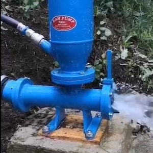 Op de afbeelding: Een blauwe waterpomp met een rood label met de tekst "RAM PUMP TURKIE" is gemonteerd op een houten platform. De pomp is aangesloten op een slang en pompt water.