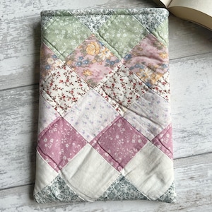 May include: A patchwork book cover in a diamond pattern. The cover is made from various floral and solid fabrics in shades of pink, green, and white. The cover is designed to protect a book, with a quilted texture.