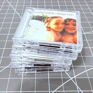 May include: A stack of clear plastic CD cases. The top case displays the album cover for "Siamese Dream" by The Smashing Pumpkins, featuring a photo of two young girls. The cases are stacked on a gray cutting mat.