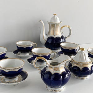 Könnte beinhalten: Vintage Teeservice mit marineblauem und goldenem Design. Das Set enthält eine Teekanne, einen Sahnekännchen, eine Zuckerdose, Untertassen und Teetassen. Die Tassen und Untertassen haben goldene Ränder. Die Teekanne hat einen weißen Deckel und Griff.