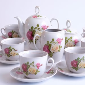 May include: A white porcelain tea set featuring a teapot, creamer, sugar bowl, and multiple teacups with saucers. Each piece is decorated with a floral pattern of pink and yellow roses and green leaves.
