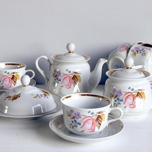 May include: A white porcelain tea set featuring a delicate floral pattern with pink tulips, blue flowers, and brown leaves. The set includes a teapot, a lidded sugar bowl, a lidded butter dish, and multiple teacups with matching saucers, all accented with gold trim.