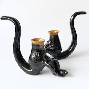 May include: Two dark green ceramic pipes with a glossy finish. Each pipe has a curved stem and a small bowl with a tan rim. The pipes are decorated with floral and leaf motifs.