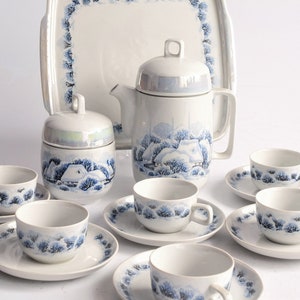 May include: A vintage white porcelain tea set with a winter landscape design. The set includes a teapot, sugar bowl, serving tray, and six teacups with saucers. The design features blue trees and houses.