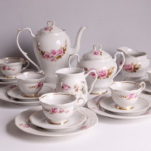 May include: A white porcelain tea set featuring pink rose floral patterns and gold edging. The set comprises a teapot, sugar bowl, creamer, cups, and saucers. The tea set is displayed on a white surface.