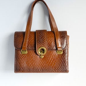 May include: A vintage brown handbag with a reptile-effect texture. It has a top handle, a flap closure with a gold-coloured clasp, and matching gold-coloured hardware. The bag is rectangular in shape, with a classic design.