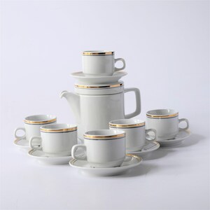 May include: A white porcelain coffee set with gold trim. The set includes a coffee pot, six cups, and saucers. The cups and pot have a black line below the gold trim. The set is arranged on a white surface.
