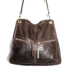 May include: A dark brown leather shoulder bag with two front zipped pockets. The bag has a curved top and a long strap with silver-coloured metal hardware. The leather has visible stitching and a slightly distressed appearance.
