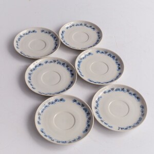 May include: Six off-white ceramic saucers with a blue floral pattern around the rim. The saucers are arranged in a circular pattern, showcasing their design. The saucers are a classic addition to any tea set.