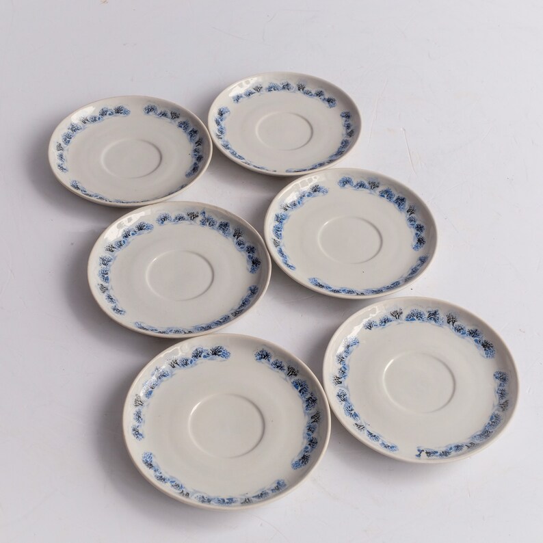 May include: Six off-white ceramic saucers with a blue floral pattern around the rim. The saucers are arranged in a circular pattern, showcasing their design. The saucers are a classic addition to any tea set.