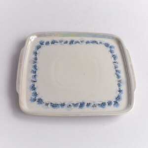 May include: A rectangular ceramic serving tray with a cream-colored surface and a border of blue floral designs. The tray has two small handles and a slightly iridescent rim.