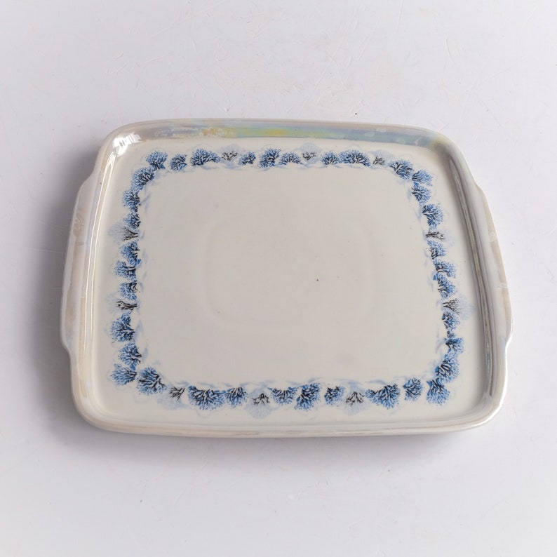 May include: A rectangular ceramic serving tray with a cream-colored surface and a border of blue floral designs. The tray has two small handles and a slightly iridescent rim.